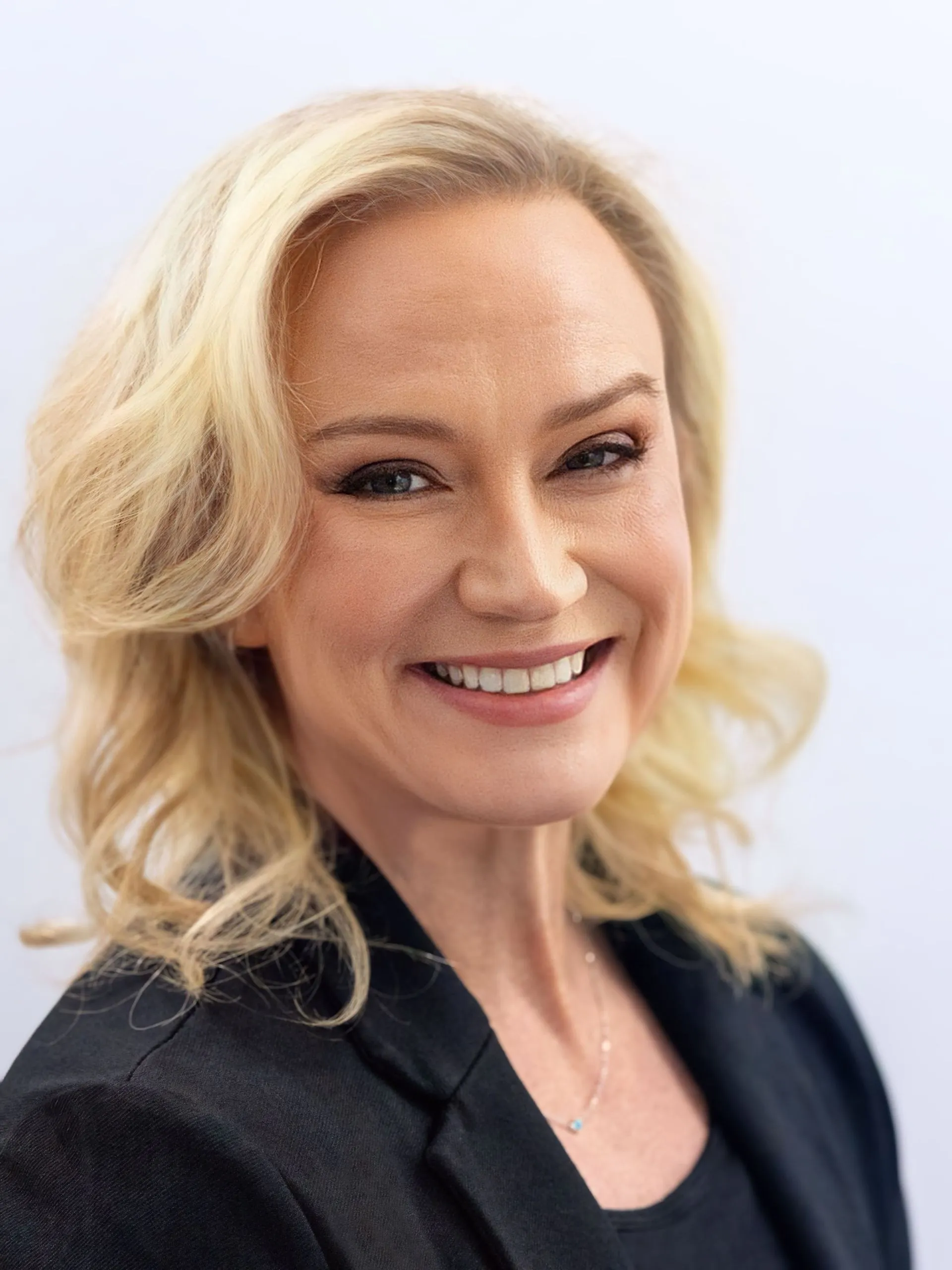 Professional headshot of a smiling blonde woman wearing a black blazer and necklace.