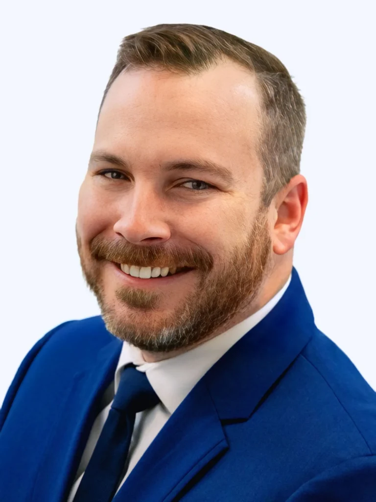 Portrait of a smiling man with a beard wearing a blue suit and tie against a light background.
