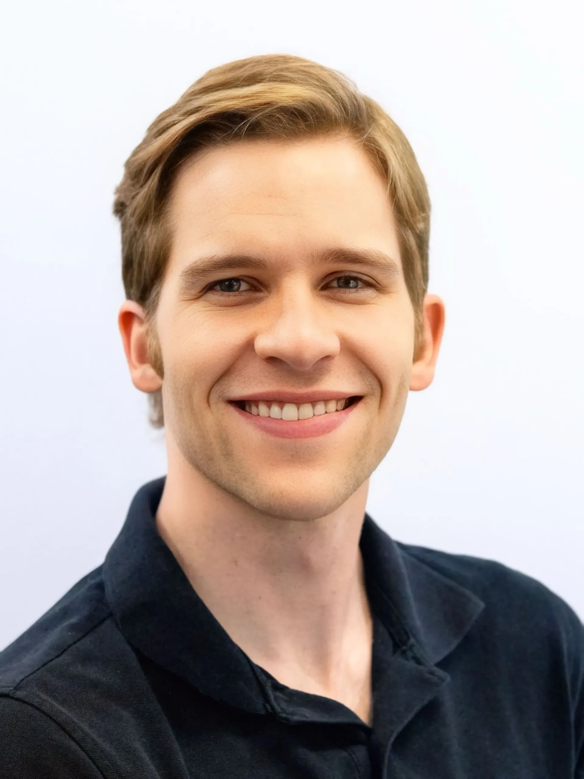 Professional headshot of a smiling young man with light brown hair wearing a black collared shirt.