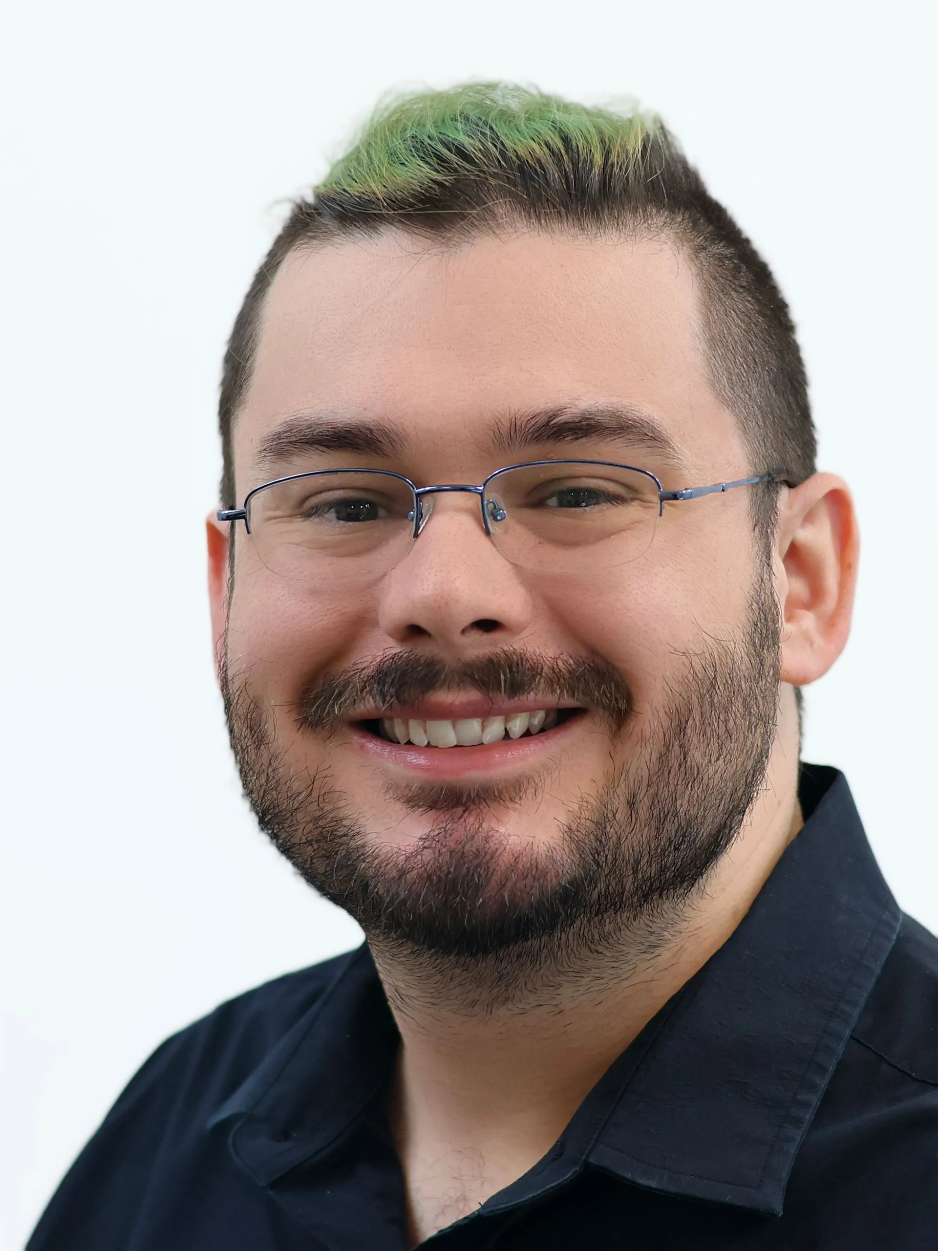 Portrait of a smiling man with short dark hair, green highlights, glasses, and a beard wearing a black shirt.