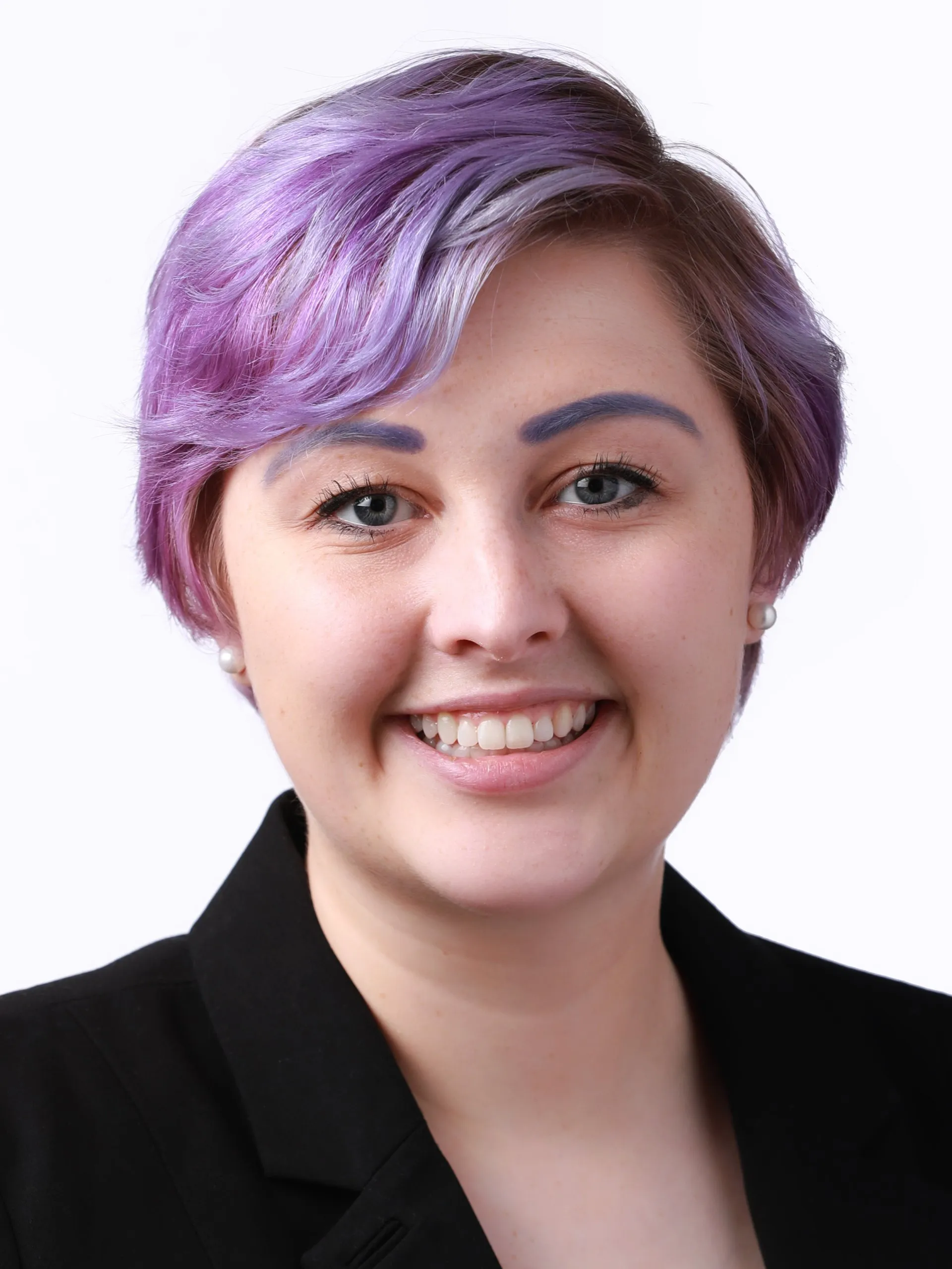 Professional headshot of a smiling woman with short purple hair and matching eyebrows, wearing a black blazer.