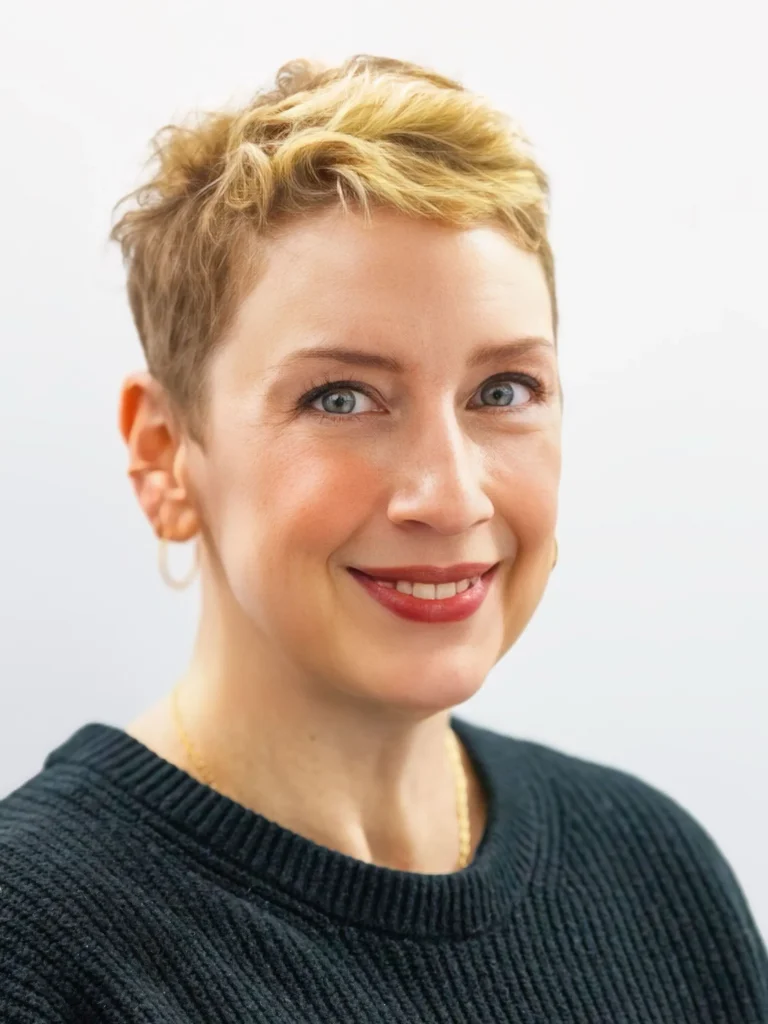 Professional portrait of a smiling woman with short blonde hair, wearing a black sweater and gold hoop earrings.