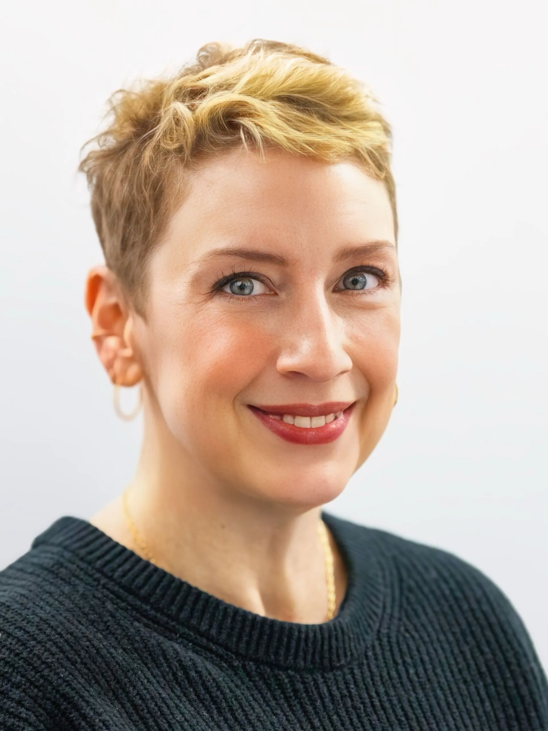 Professional portrait of a smiling woman with short blonde hair, wearing a black sweater and gold hoop earrings.