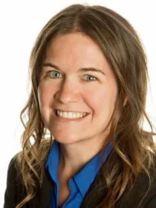 Professional headshot of a smiling woman with light brown hair wearing a blue shirt and black blazer.