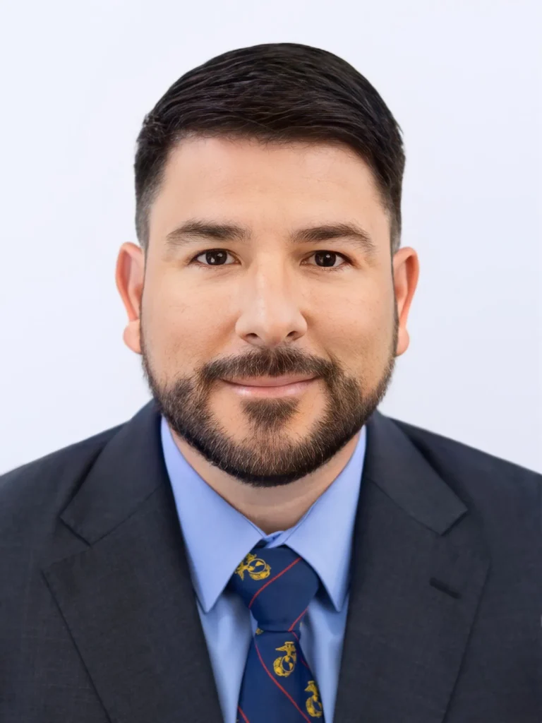 Professional headshot of a man with dark hair and beard wearing a dark suit, blue shirt, and Marine Corps-themed tie.