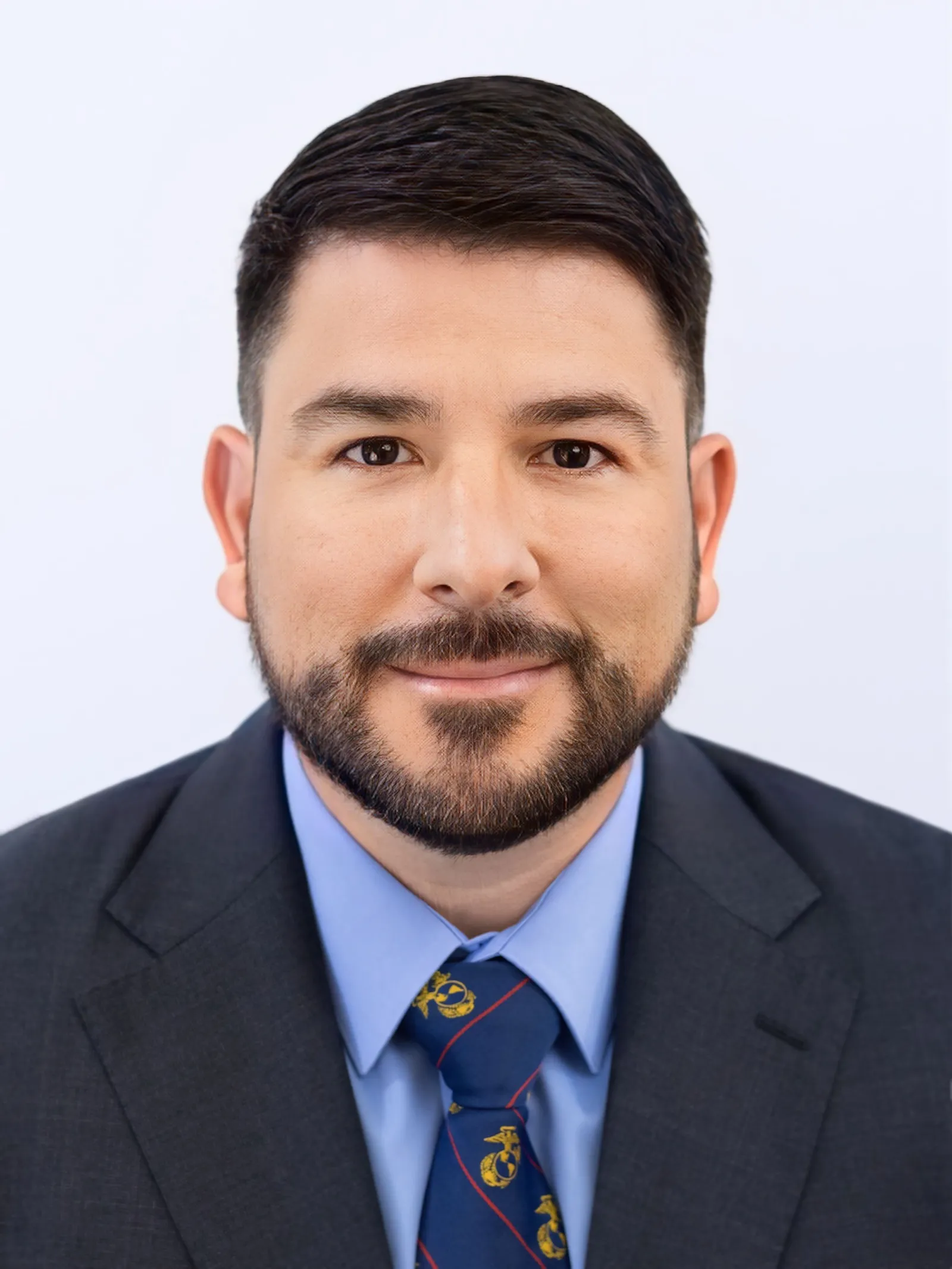 Professional headshot of a man with dark hair and beard wearing a dark suit, blue shirt, and Marine Corps-themed tie.