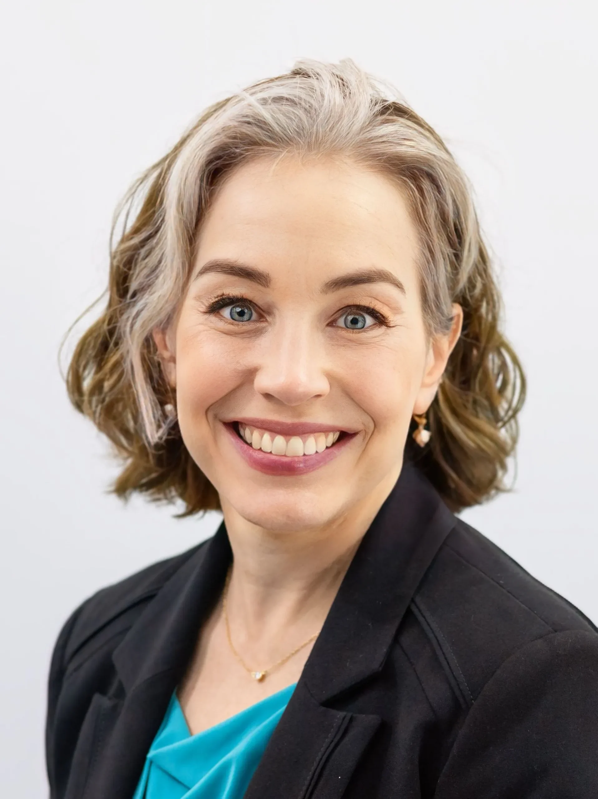 Professional headshot of a smiling woman with short gray and brown hair, wearing a black blazer and teal blouse.