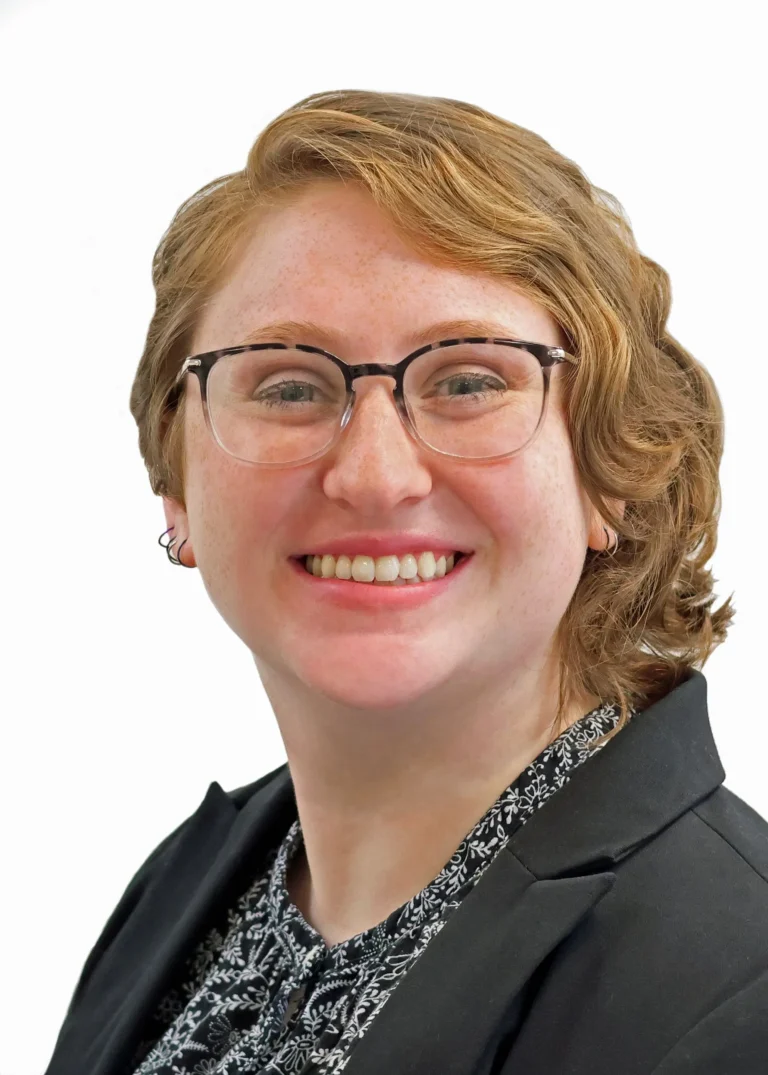 Professional portrait of a smiling woman with short red hair, glasses, black blazer, and patterned blouse.