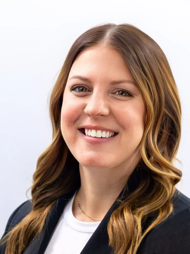 Professional headshot of a smiling woman with wavy brown hair, wearing a black blazer and white top.