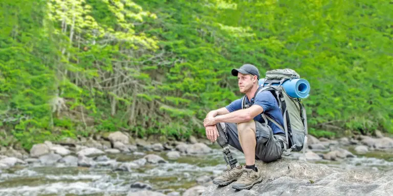 Man with backpack and prosthetic leg sitting on rock by river in forest.
