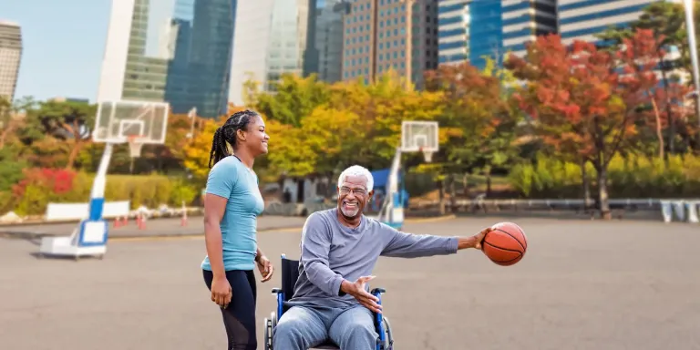 An elderly man in a wheelchair holding a basketball, smiling at a standing woman on an outdoor court.