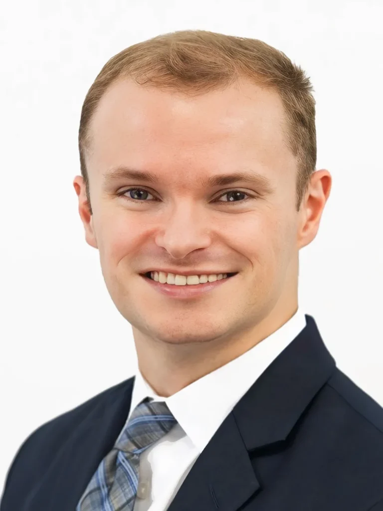 Professional headshot of a smiling young man with light brown hair, wearing a dark suit and blue patterned tie.